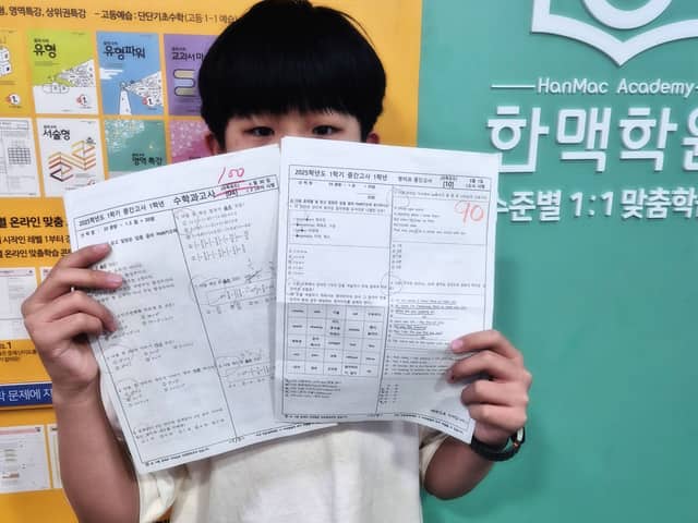 학원 로고 벽 앞에서 90점과 100점 시험지를 들어 보이는 학생
